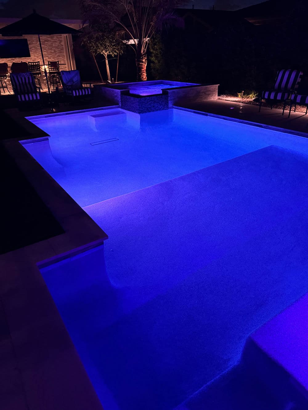 Illuminated swimming pool at night with blue lighting and modern outdoor furniture.
