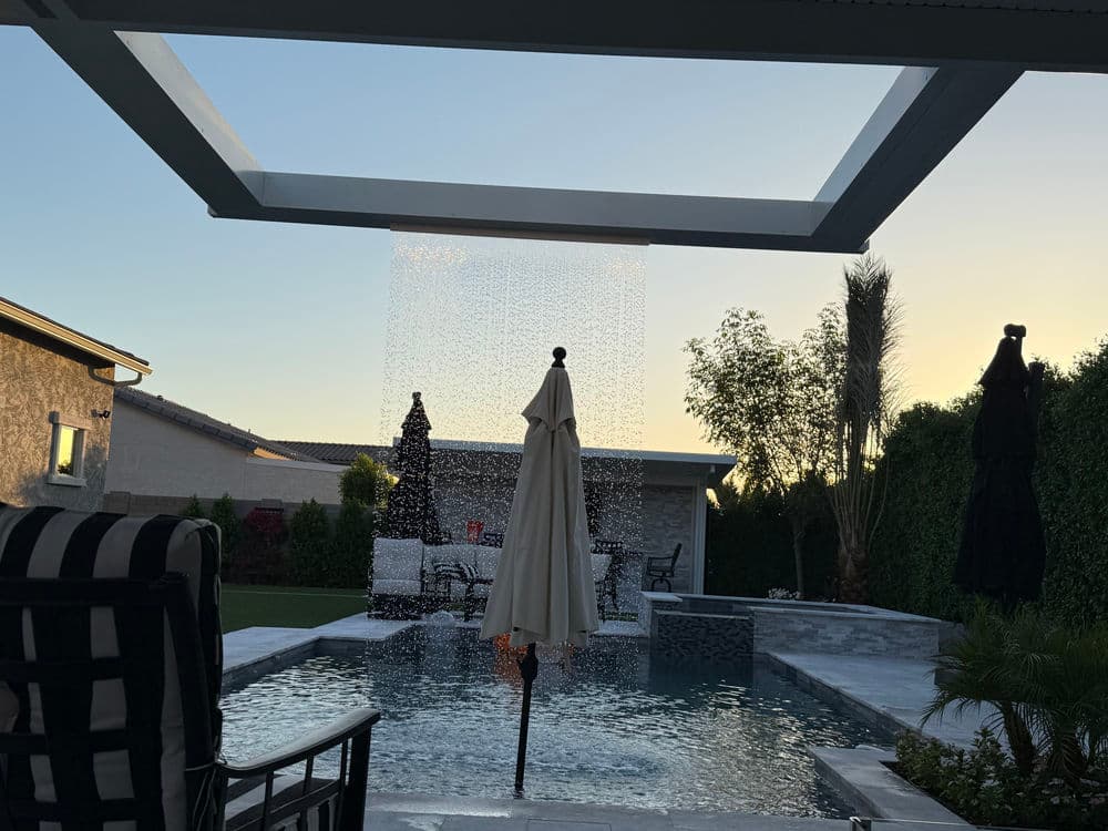 Modern backyard oasis with a pool, waterfall feature, and patio umbrella at sunset.