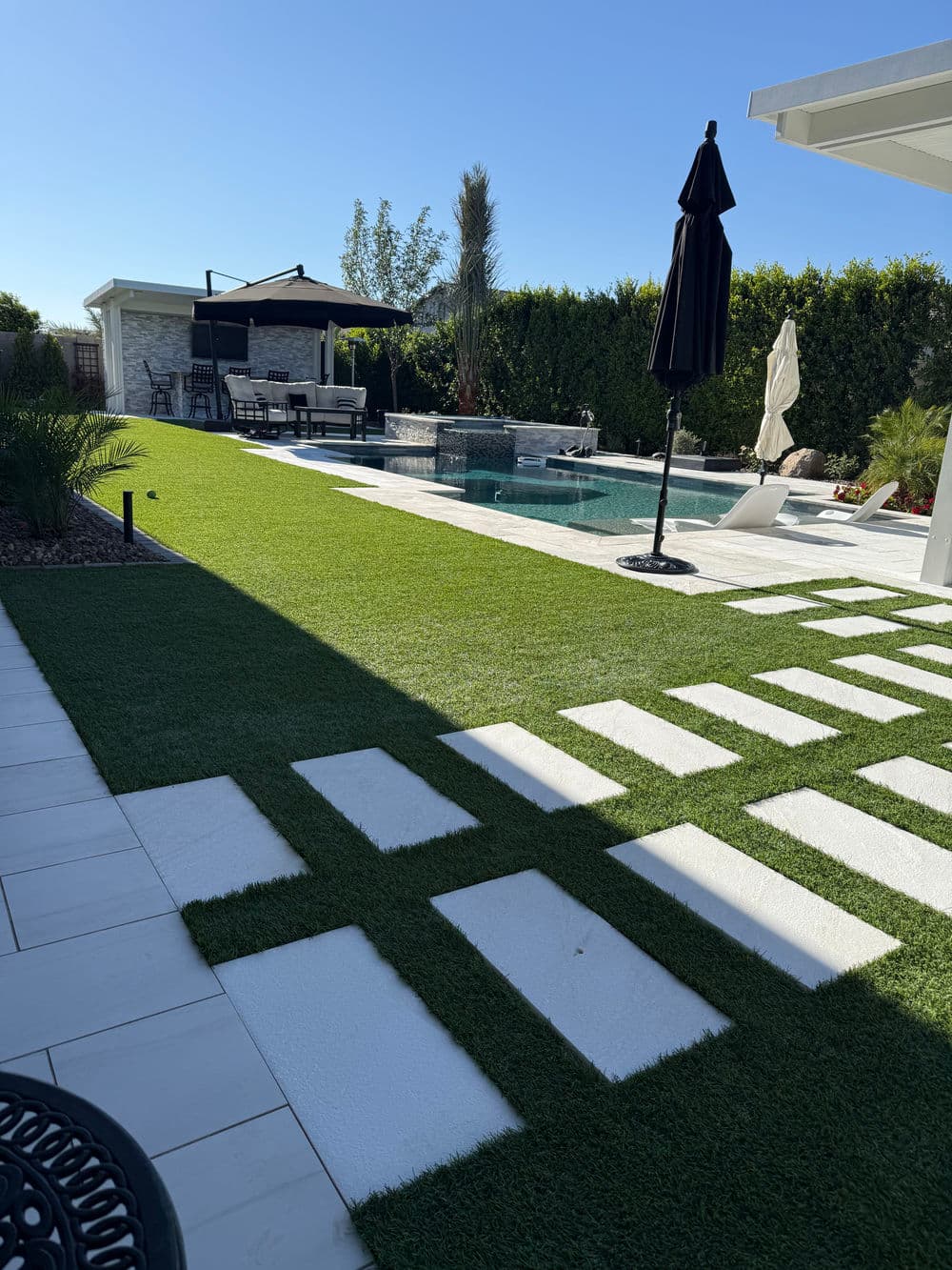 Modern backyard with a pool, artificial grass, and patio furniture under clear blue sky.