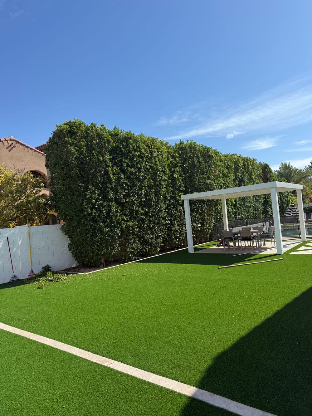 Lush artificial grass yard with green hedge and outdoor dining area under a pergola.