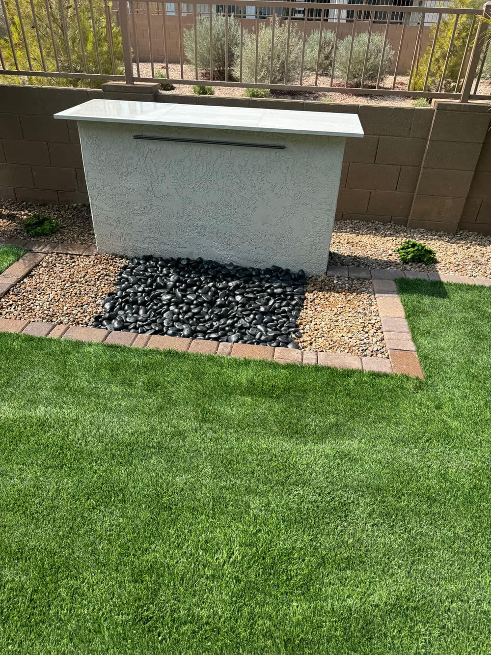 Modern outdoor water feature with black pebbles and lush green grass in backyard setting.