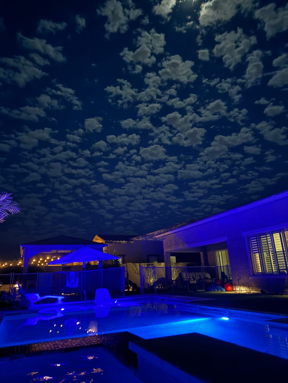 Night view of a pool with blue lighting, illuminated clouds, and a modern home.