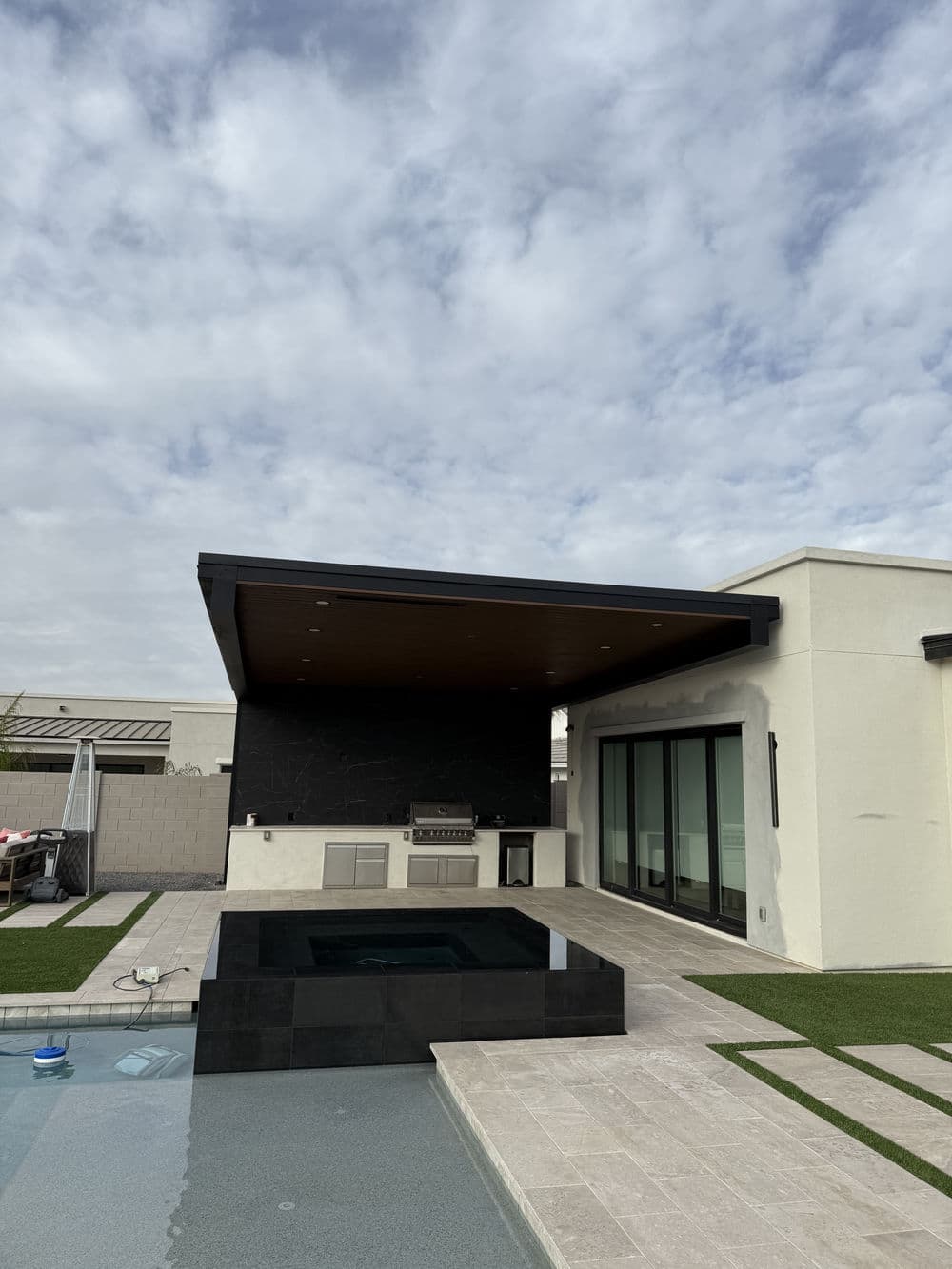 Modern backyard with a pool, outdoor kitchen, and stylish covered patio under a cloudy sky.