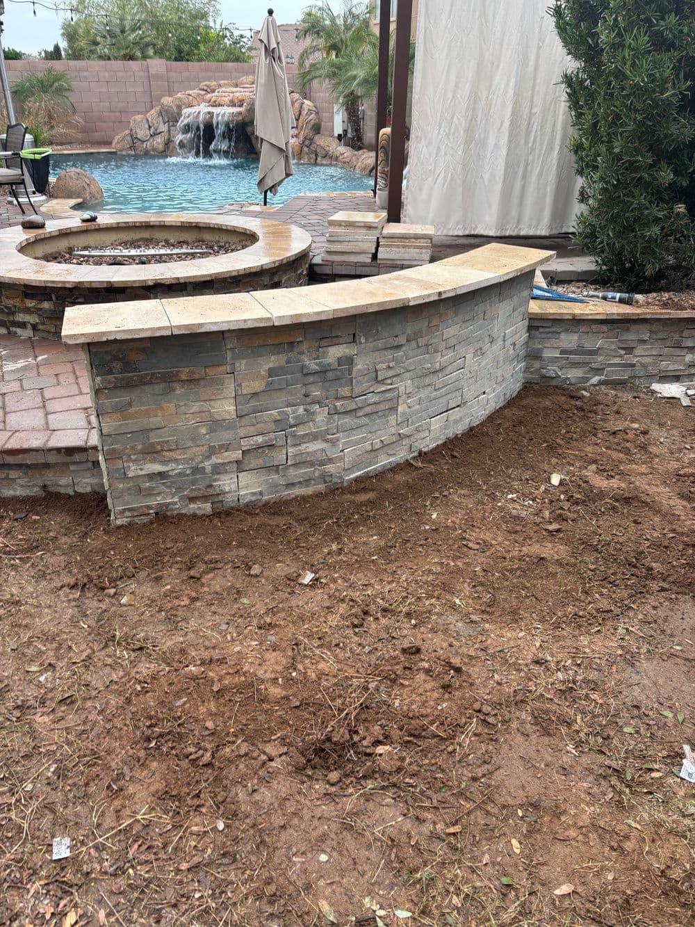 Stone seating wall by pool with waterfall, surrounded by dirt and landscaping work in progress.