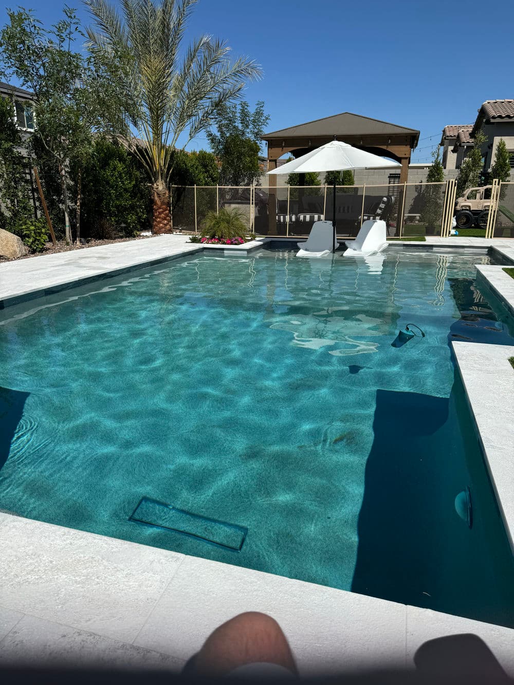 Backyard swimming pool with lounge chairs, palm trees, and a shaded seating area.
