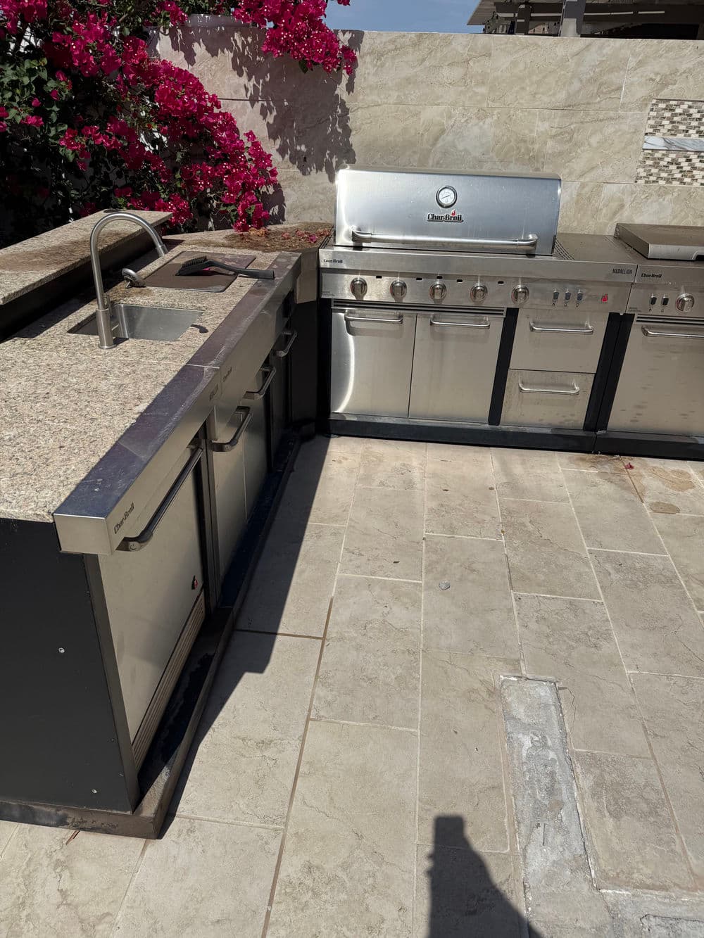 Outdoor kitchen with stainless steel appliances and granite countertop, surrounded by flowering plants.