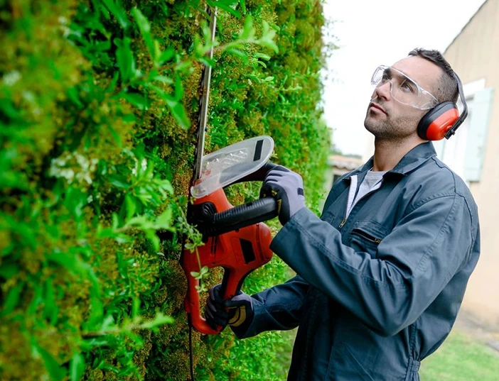 A man with goggles and gloves on using an electric saw to cut the branches of a tree.